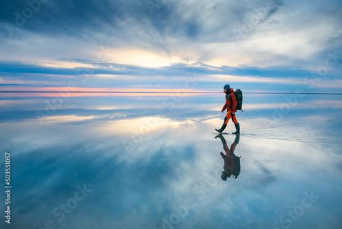 Man traveler with backpack walking on the salt lake at sunset. Sky with clouds are reflected in the mirror water surface. Travel and adventure concept