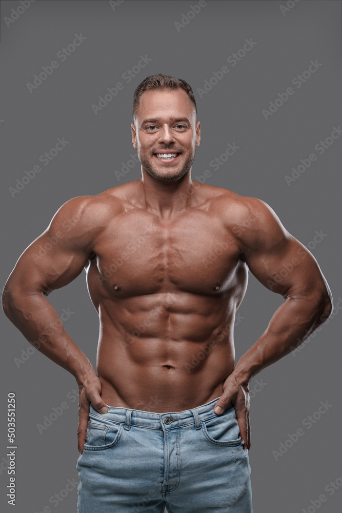 Studio shot of handsome guy with muscular build dressed in jeans posing against grey background.
