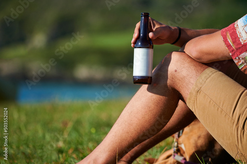 Crop male sitting with bottle of beer on grassy lawn near the sea and enjoying sunny day in summer