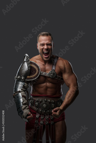 Shot of brutal roman gladiator dressed in light armor screaming at camera against gray background.