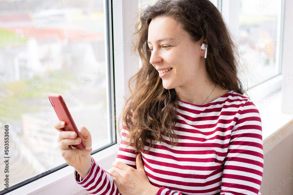 Pretty brunette woman using smartphone standing near window at home. Checking social media, chatting with friend, reading news, watching online webinar, shopping, browsing website, ordering delivery Pretty brunette woman using smartphone standing near window at home. Checking social media, chatting with friend, reading news, watching online webinar, shopping, browsing website, ordering delivery