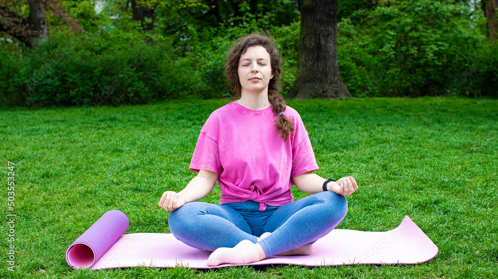 Calm young woman with closed eyes sitting in lotus pose, practicing yoga breathing techniques on green grass in garden outdoor,wearing pink t-shirt.Meditation and pranayama.Mental health care concept