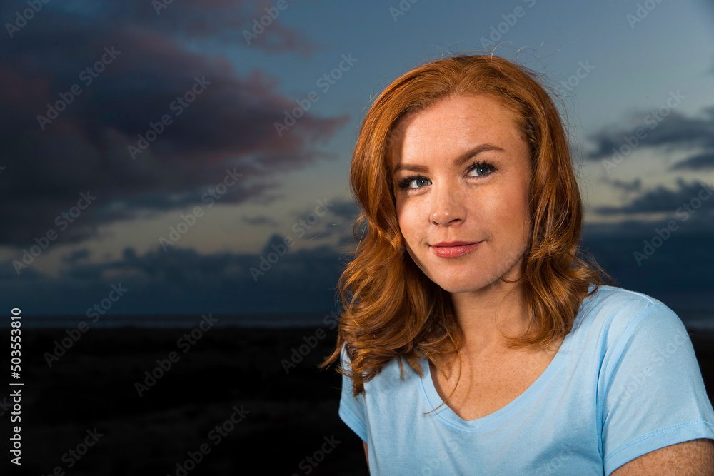 Pretty redhead woman with freckles wearing blue shirt outside in the evening at ocean beach with a view.