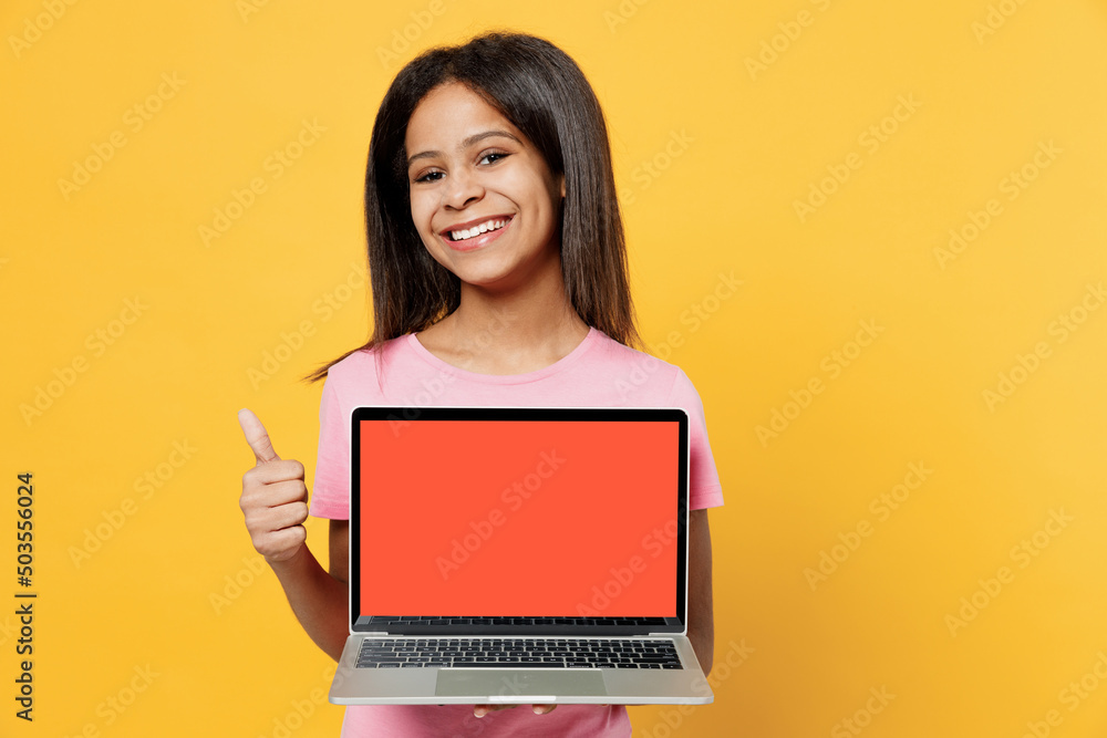 Naklejka premium Little kid girl of African American ethnicity 12-13 years old in pink t-shirt hold use work on laptop pc computer with blank screen workspace area show thumb up isolated on plain yellow background