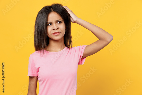 Little puzzled sad kid girl of African American ethnicity 12-13 years old in pink t-shirt look aside scratch hold head isolated on plain yellow background studio portrait. Childhood lifestyle concept.