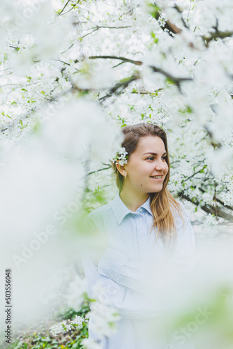 Young pretty woman enjoys standing near flowering spring tree. A girl wearing beige hat and white dress smiles among blooming apple trees. Spring season concept © Дмитрий Ткачук