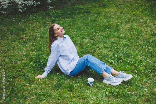 Young hipster woman in denim jacket is resting on green lawn in park, drinking coffee, relaxing outdoors. Lifestyle, coffee break. Selective focus on female hands holding disposable cup of coffee. © Дмитрий Ткачук
