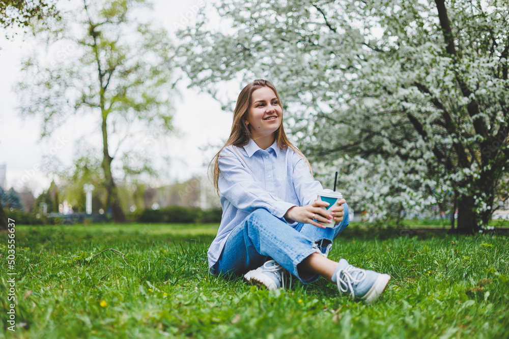 Young hipster woman in denim jacket is resting on green lawn in park, drinking coffee, relaxing outdoors. Lifestyle, coffee break. Selective focus on female hands holding disposable cup of coffee. Young hipster woman in denim jacket is resting on green lawn in park, drinking coffee, relaxing outdoors. Lifestyle, coffee break. Selective focus on female hands holding disposable cup of coffee.
