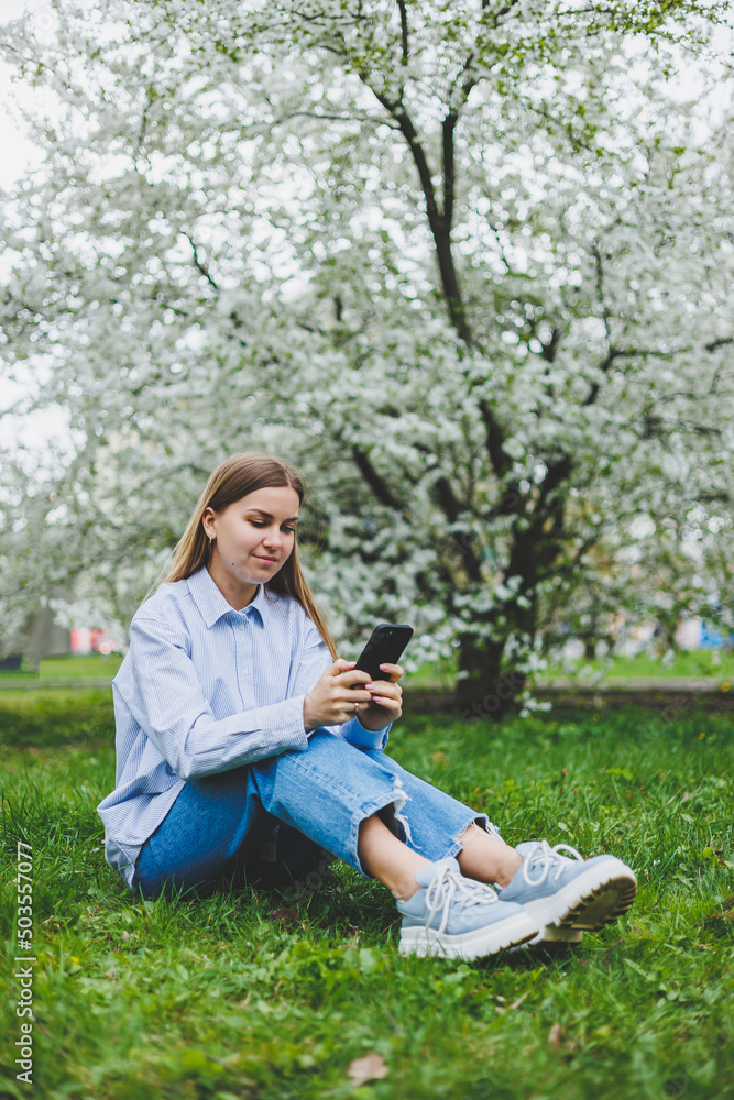 Happy woman walking using smart phone in a city street. Business woman with sunglasses uses mobile phone outdoors under blooming tree, girl typing message, chatting, scrolling web page, look at camera Happy woman walking using smart phone in a city street. Business woman with sunglasses uses mobile phone outdoors under blooming tree, girl typing message, chatting, scrolling web page, look at camera