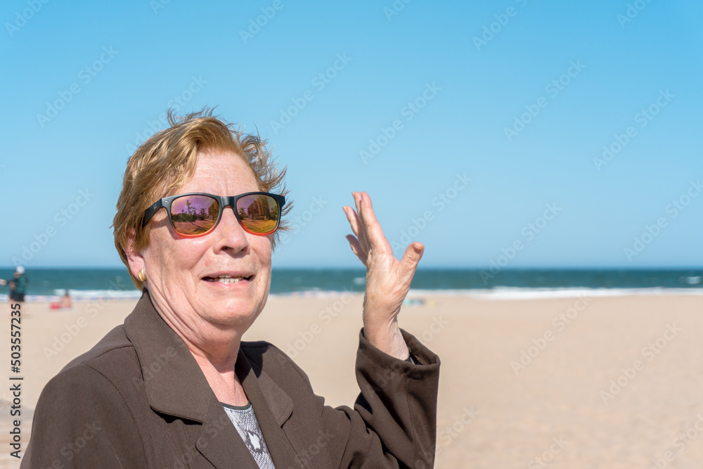 Funny older woman, with colorful sunglasses, waving on a sunny day at the beach.
