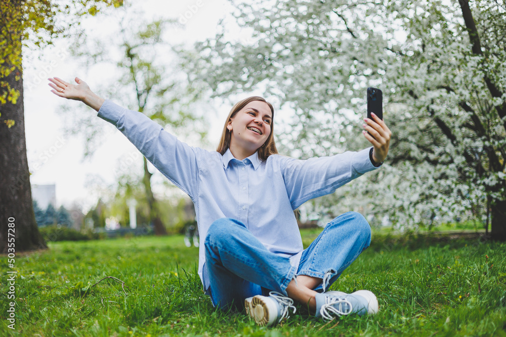 Fototapeta premium Happy woman walking using smart phone in a city street. Business woman with sunglasses uses mobile phone outdoors under blooming tree, girl typing message, chatting, scrolling web page, look at camera