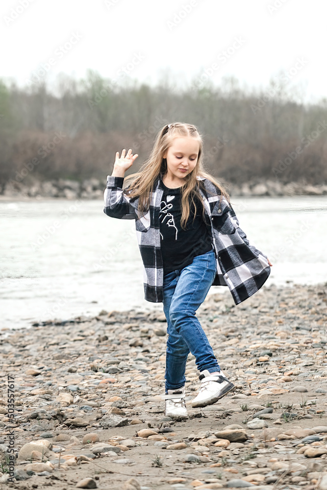 a beautiful girl of eight years old with long blond hair in a fashionable plaid shirt and jeans has fun and dances on a walk by the river