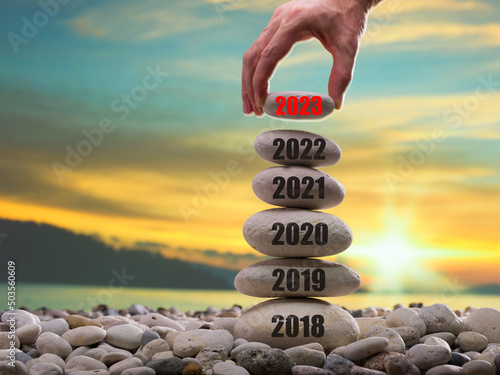 Happy new year 2023 concept. Entering the year 2023. Years ( 2018-2019-2020-2021-2022 ) written on the rising stone pile. Man hand adding stone to tower. Background is blurred sunset sky