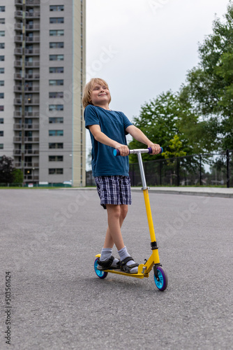 Child riding scooter. Smiling boy spending time outside in summer. Kid having fun near an apartment building in the city.