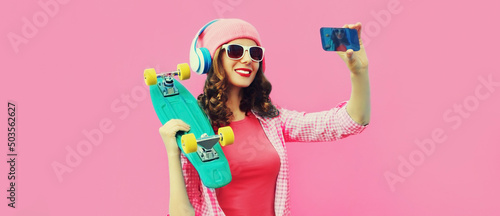 Summer colorful portrait of stylish modern young woman taking a selfie by smartphone with skateboard and headphones on pink background