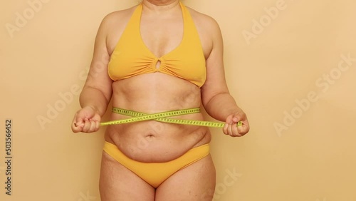 Unrecognizable fat overweight woman standing in yellow bra, bikini, showing excess belly, tightening waist with tape on beige background. Body positive, cellulite, obesity, weight control, measuring.