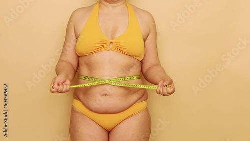 Unrecognizable fat overweight woman standing in yellow bra, bikini, showing excess belly, tightening waist with tape on beige background. Body positive, cellulite, obesity, weight control, measuring. photo