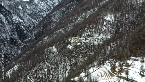 Flying above the swiss alps in the winter.  A small, old church sits on the side of the mountain.  Bare trees and the snow show the season.  Grey, snowy skies over the mountains.  photo