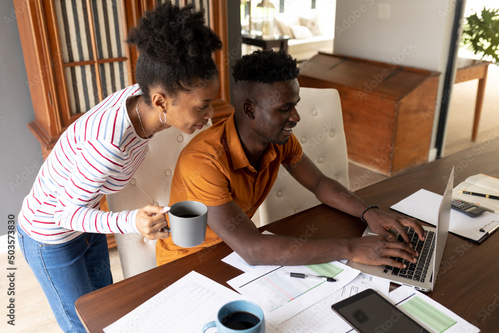 Smiling african american young woman with coffee mug standing by boyfriend using laptop on table Smiling african american young woman with coffee mug standing by boyfriend using laptop on table