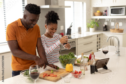 Smiling african american young couple with wine and digital tablet on kitchen island preparing food