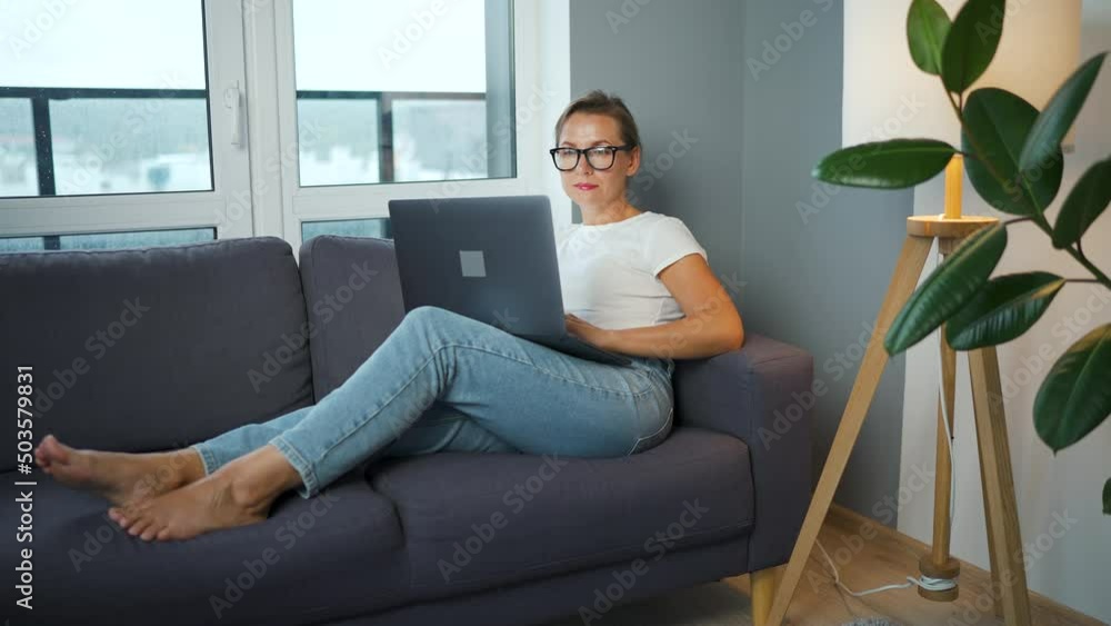 Woman with glasses is sitting on the couch and working on a laptop. Concept of remote work. Woman with glasses is sitting on the couch and working on a laptop. Concept of remote work.