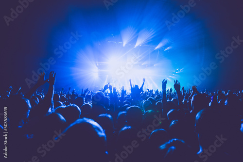 A crowded concert hall with scene stage lights in blue tones, rock show performance, with people silhouette, on dance floor air during a concert festival photo