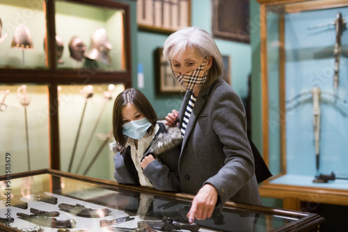 Interested preteen girl and elderly woman wearing protective face masks viewing antique handguns displayed in glass showcase in history museum. Concept of precautions in coronavirus pandemic