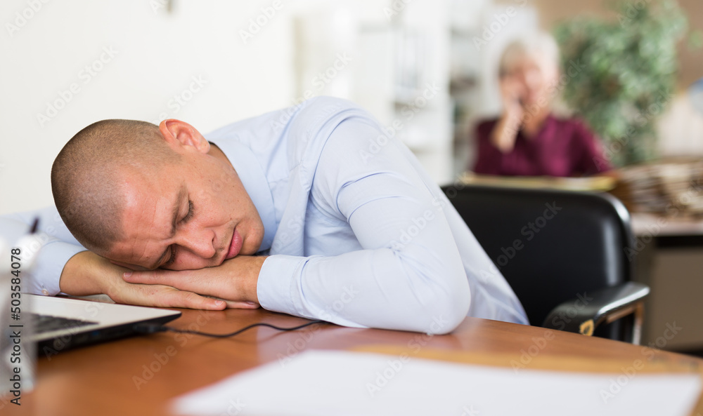 Fototapeta premium Exhausted overworked businessman sleeping on table with laptop and documents, resting his head on hands. Daily work and fatigue concept
