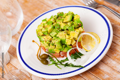Delicate raw salmon tartare with diced avocado garnished with sesame seeds, greens, lemon and capers ..