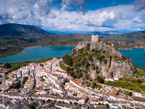 Aerial view of fortified castle on top of crag and white residential houses at bottom of hill in Zahara de la Sierra on background of turquoise lake, Spain
