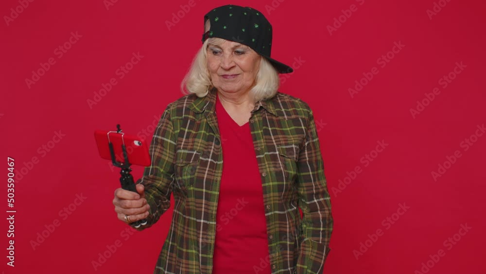Senior woman in shirt traveler blogger taking selfie on mobile phone selfie stick communicating video call online with subscribers. Elderly old grandmother isolated alone on red studio wall background Senior woman in shirt traveler blogger taking selfie on mobile phone selfie stick communicating video call online with subscribers. Elderly old grandmother isolated alone on red studio wall background