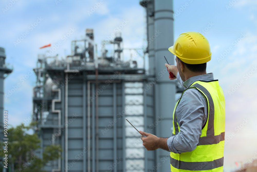 engineer at work. Engineer wearing PPE uniform and helmet looking detail tablet on hand with power plant on background. engineer at work. Engineer wearing PPE uniform and helmet looking detail tablet on hand with power plant on background.
