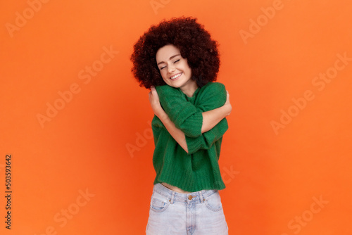 Fototapeta Woman with Afro hairstyle wearing green casual sweater hugging herself and smiling, feeling comfortable and fulfilled, narcissistic egoistic person