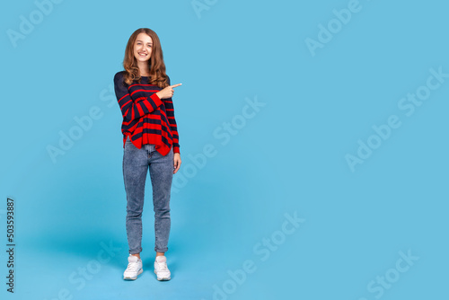 Full length portrait of smiling woman wearing sweater, pointing finger aside, showing copy space for commercial text, blank wall with idea presentation. Indoor studio shot isolated on blue background.