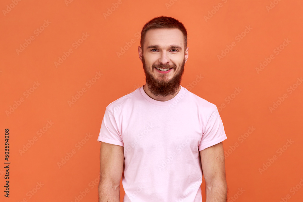 Portrait of happy bearded man looking at camera and smiling, expressing positive emotions, being in good mood, wearing pink T-shirt. Indoor studio shot isolated on orange background. Portrait of happy bearded man looking at camera and smiling, expressing positive emotions, being in good mood, wearing pink T-shirt. Indoor studio shot isolated on orange background.