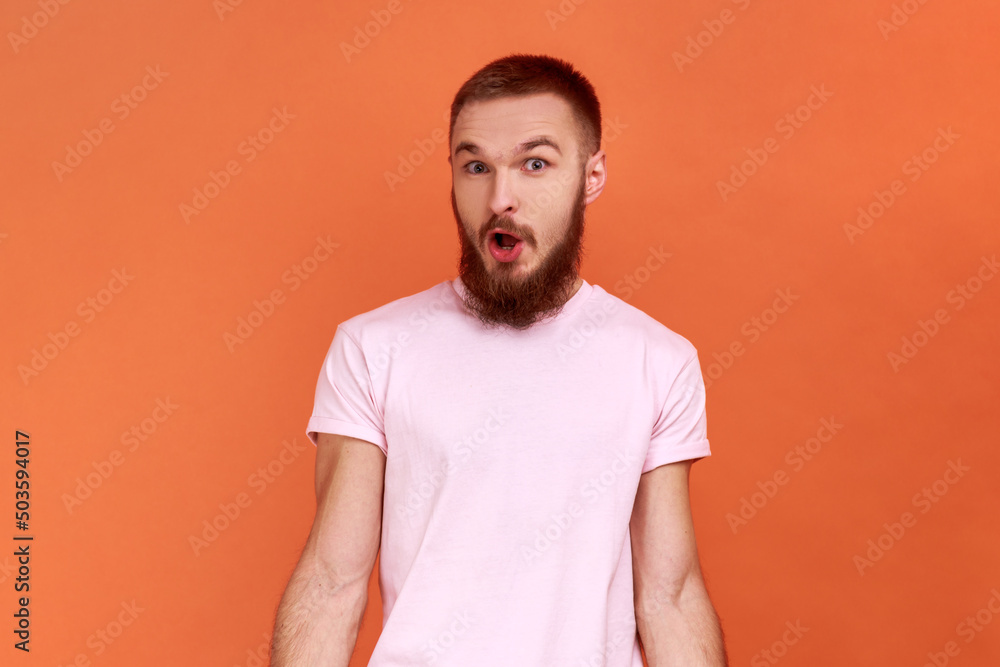 Portrait of amazed bearded man standing with mouth open in surprise, has shocked expression, hears unbelievable news, wearing pink T-shirt. Indoor studio shot isolated on orange background. Portrait of amazed bearded man standing with mouth open in surprise, has shocked expression, hears unbelievable news, wearing pink T-shirt. Indoor studio shot isolated on orange background.