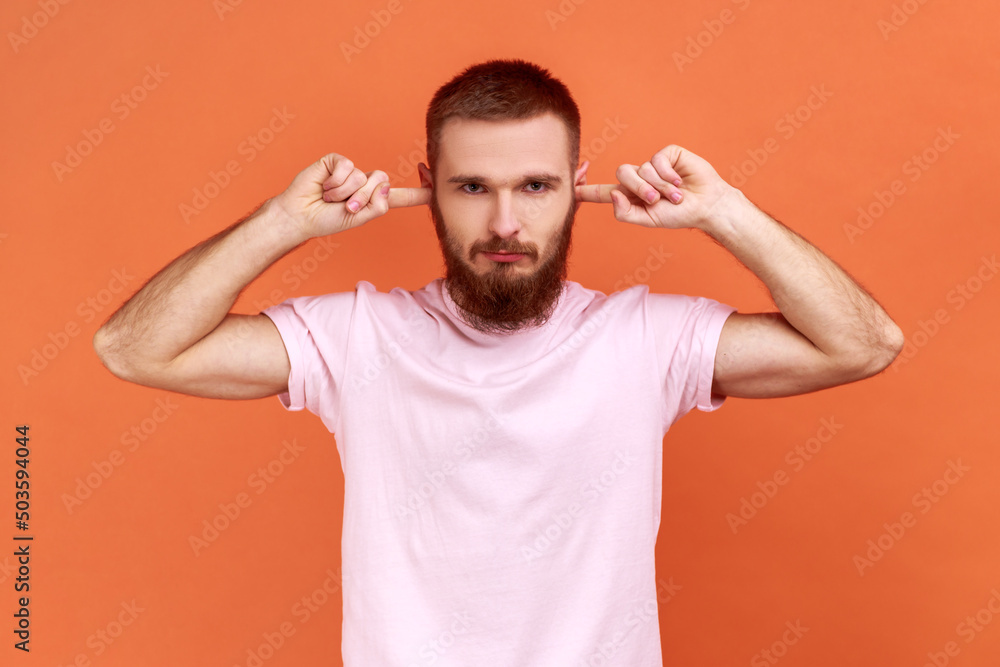 Portrait of bearded man closing ears with fingers seriously looking at camera, tired of irritated noise, wants silence, wearing pink T-shirt. Indoor studio shot isolated on orange background. Portrait of bearded man closing ears with fingers seriously looking at camera, tired of irritated noise, wants silence, wearing pink T-shirt. Indoor studio shot isolated on orange background.