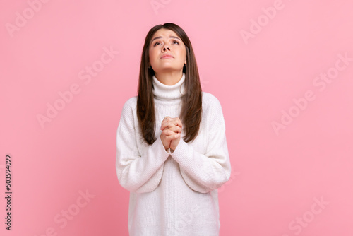 Fototapeta Young adult attractive brunette girl looking up with hands in prayer, sincere saying please, apology, wearing white casual style sweater