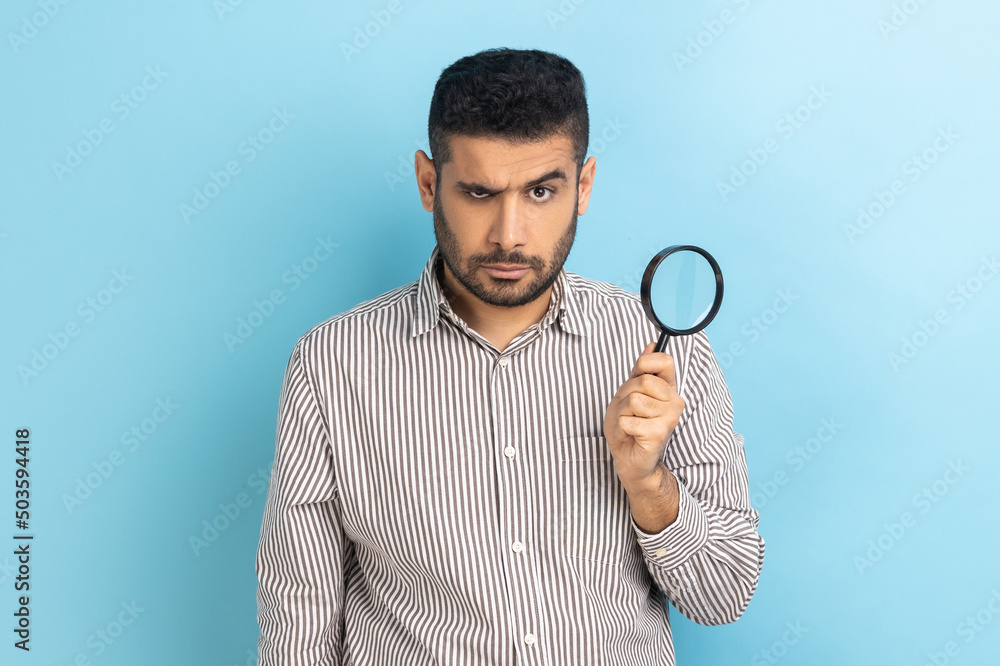 Portrait of serious confident businessman holding in hand magnifying glass and looking at camera, spying, wearing striped shirt. Indoor studio shot isolated on blue background. Portrait of serious confident businessman holding in hand magnifying glass and looking at camera, spying, wearing striped shirt. Indoor studio shot isolated on blue background.