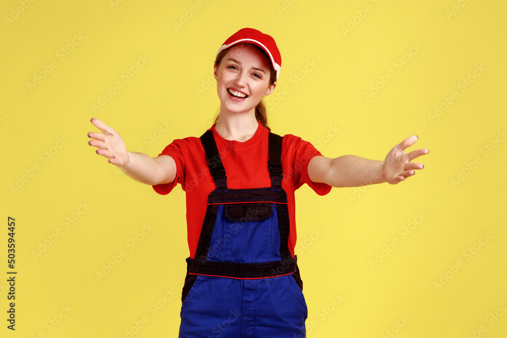 Happy smiling handy woman standing with outstretched hands, inviting to embrace, come into my arms, wearing overalls and red cap. Indoor studio shot isolated on yellow background. Happy smiling handy woman standing with outstretched hands, inviting to embrace, come into my arms, wearing overalls and red cap. Indoor studio shot isolated on yellow background.