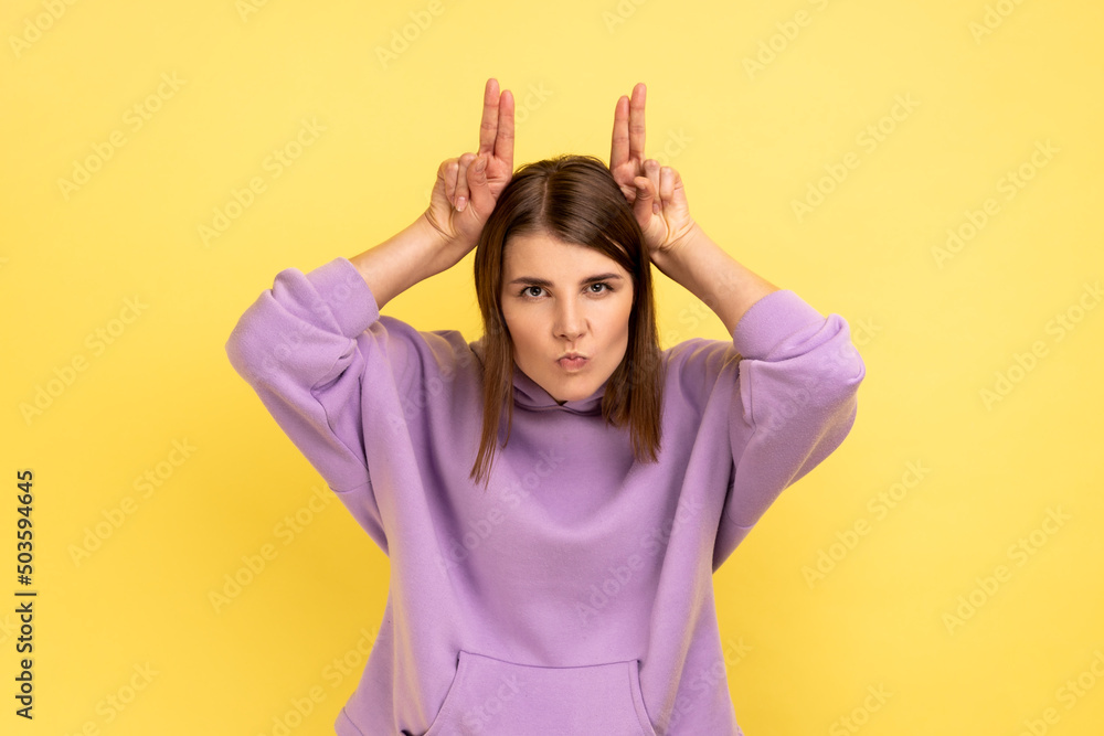Portrait of aggressive woman holding fingers above head showing horns, arrogant and stubborn, ready to attack, wearing purple hoodie. Indoor studio shot isolated on yellow background.