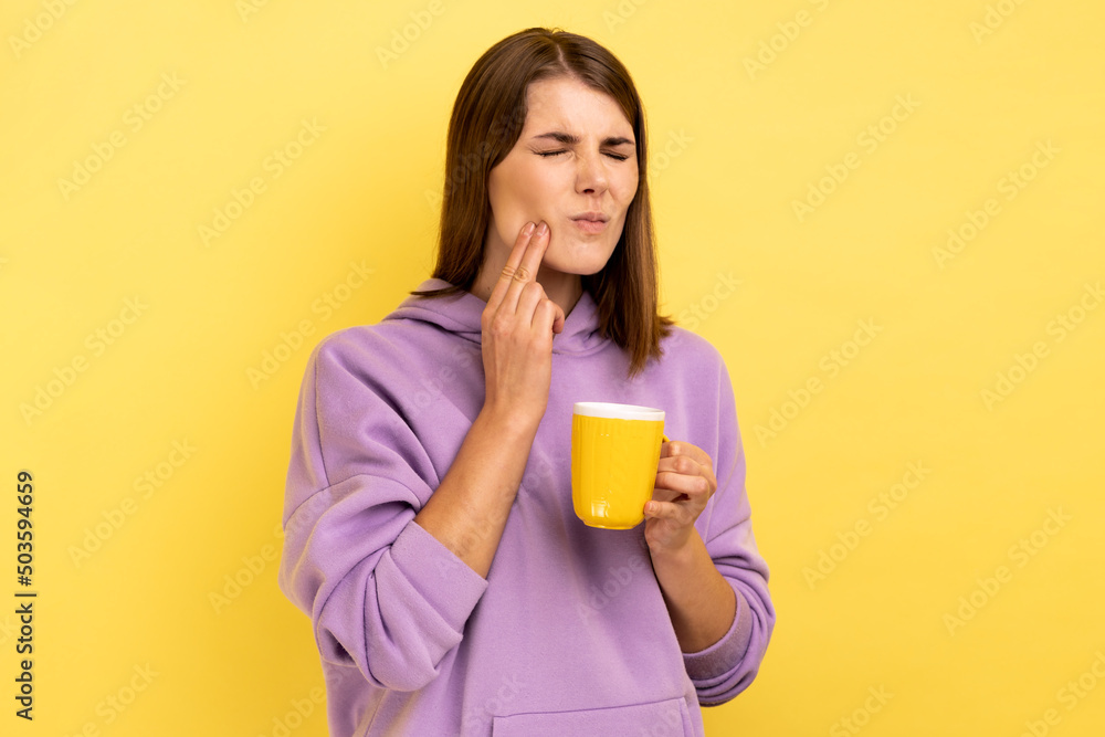 Portrait of sad sick unhealthy woman has teeth pain after drinking hot or cold beverage, dental injury, wearing purple hoodie. Indoor studio shot isolated on yellow background. Portrait of sad sick unhealthy woman has teeth pain after drinking hot or cold beverage, dental injury, wearing purple hoodie. Indoor studio shot isolated on yellow background.