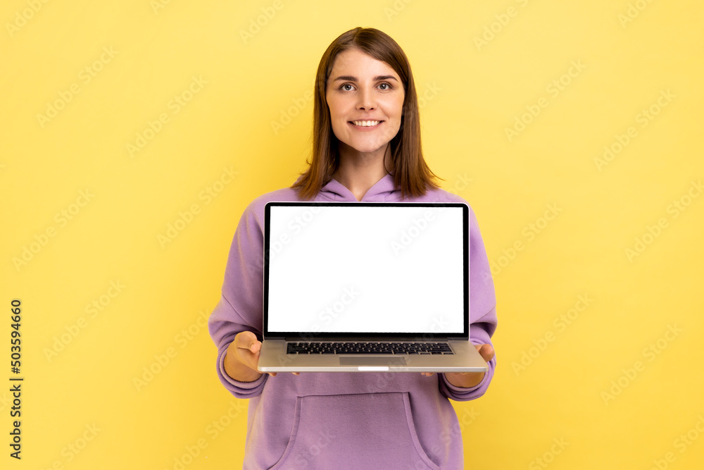Positive optimistic woman standing showing laptop with white empty screen for promotion, looking at camera with smile, wearing purple hoodie. Indoor studio shot isolated on yellow background. Positive optimistic woman standing showing laptop with white empty screen for promotion, looking at camera with smile, wearing purple hoodie. Indoor studio shot isolated on yellow background.