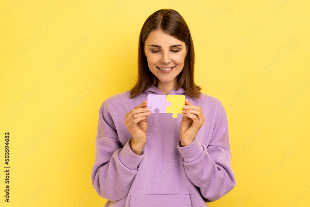 Portrait of happy smiling concentrated attractive woman holding yellow and purple puzzle pieces, solving task, wearing hoodie. Indoor studio shot isolated on yellow background. Portrait of happy smiling concentrated attractive woman holding yellow and purple puzzle pieces, solving task, wearing hoodie. Indoor studio shot isolated on yellow background.