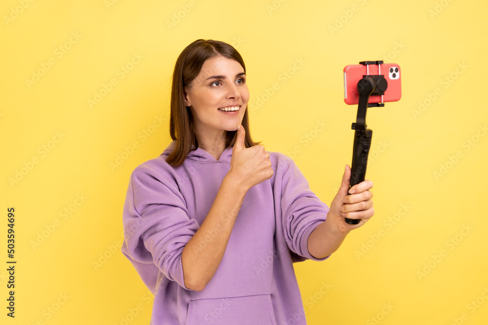 Portrait of optimistic woman using cell phone and steadicam for broadcasting livestream, showing thumb up, approved gesture, wearing purple hoodie. Indoor studio shot isolated on yellow background. Portrait of optimistic woman using cell phone and steadicam for broadcasting livestream, showing thumb up, approved gesture, wearing purple hoodie. Indoor studio shot isolated on yellow background.