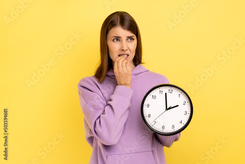 Portrait of impatient nervous attractive woman biting her nails and holding big wall clock, deadline, looking away, wearing purple hoodie. Indoor studio shot isolated on yellow background.