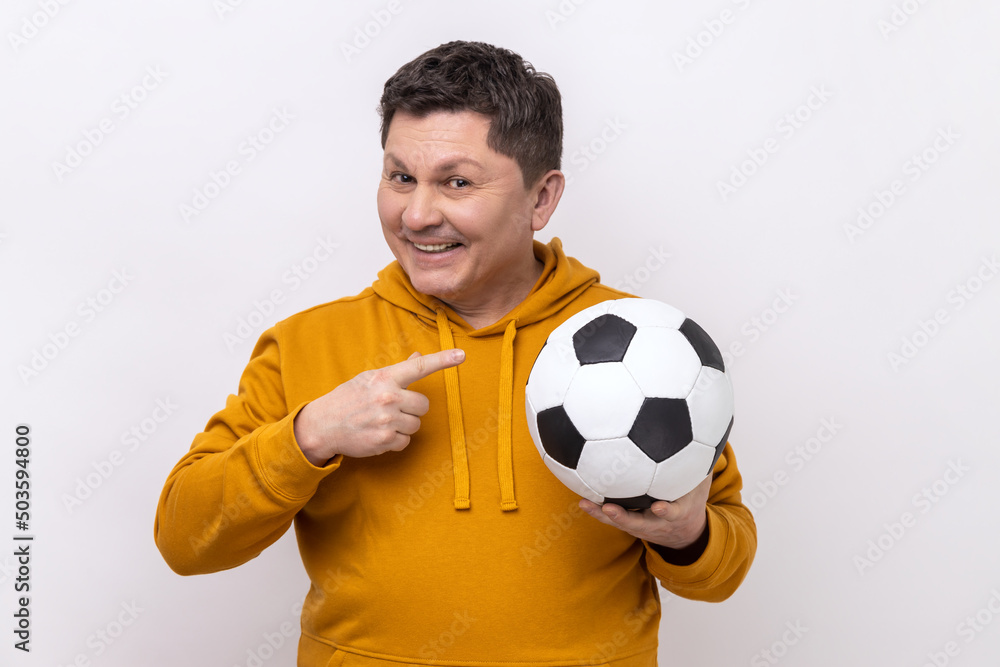 Handsome middle aged man pointing finger at soccer ball on his hand with smiling positive expression, football fan, wearing urban style hoodie. Indoor studio shot isolated on white background. Handsome middle aged man pointing finger at soccer ball on his hand with smiling positive expression, football fan, wearing urban style hoodie. Indoor studio shot isolated on white background.