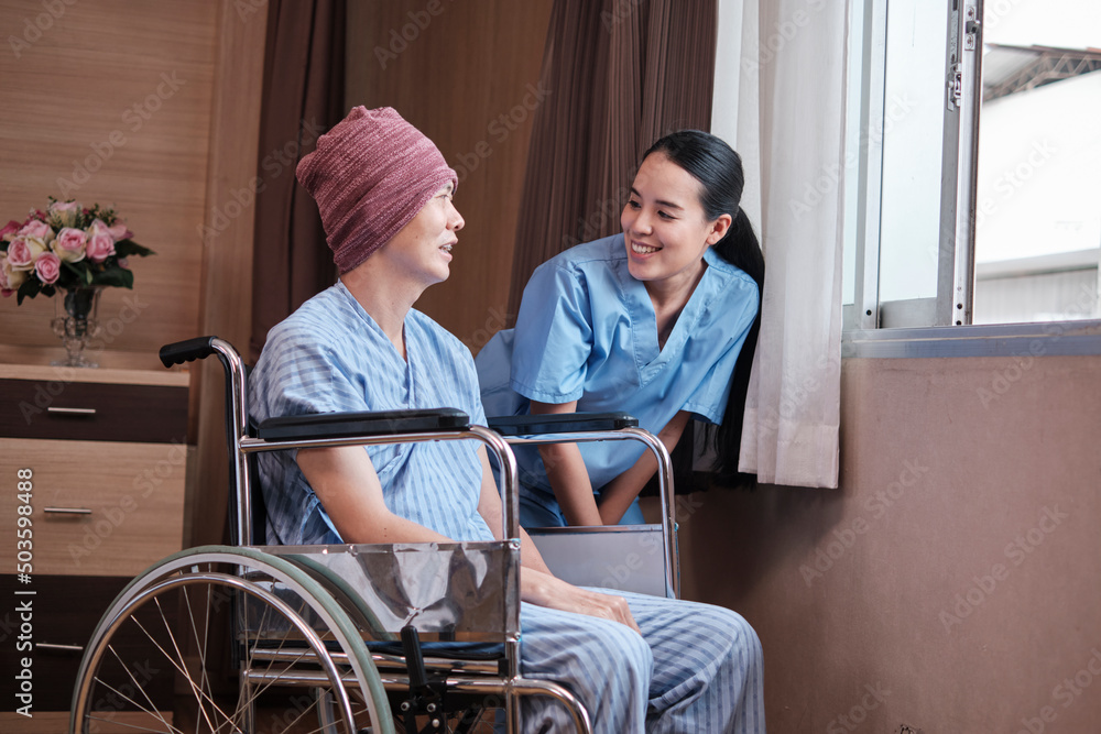 Uniformed young Asian female therapy doctor encouraging wheelchair male patient at window to support and motivate recovery, cancer illness after chemo medical treatment in hospital inpatient room.