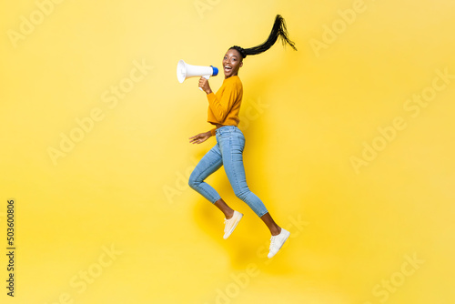 Portrait of young African American woman holding megaphone jumping in isolated yellow studio background