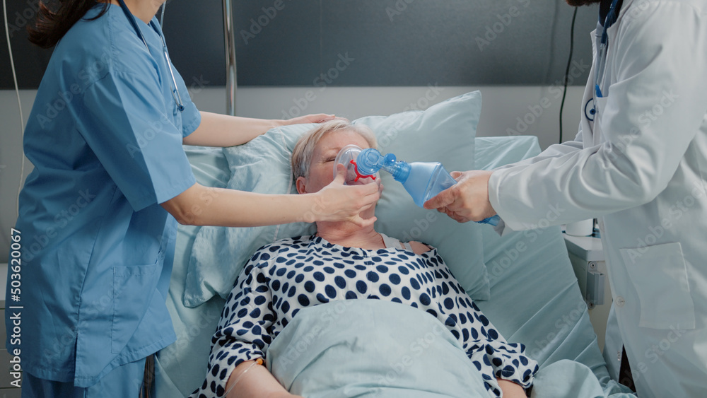 Doctor and nurse using oxygen tube for hyperventilating woman in bed. Medical specialists with tool and heart rate monitor for reanimation and respiratory problem of senior patient. - obrazy, fototapety, plakaty 